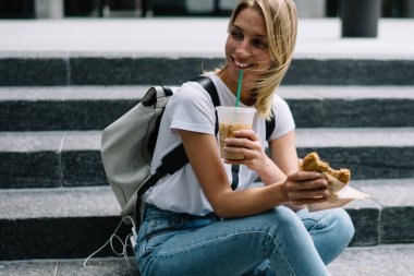 Hippi kız sokak gıda öğle yemeği yeme keyfini merdivenlerde oturan gülümsüyor, kentsel ayarları yürüyüş sırasında molası yemek tatma ve buz kahve içme mutlu kadın traveler oturan