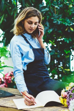 Sera, çiçek dükkanında çalışan buket kompozisyon hücresel tartışırken detayları konuşurken pazarlamacı içinde okul sırası oturan cep telefonu konuşma sırasında istemci sipariş belirterek kadın bahçıvan