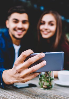 Seçici odak smartphone cep telefonu ile selfie için resim paylaşımı için cep telefonunuzun ağlarda yapma neşeli erkek ve dişi hipsters poz kafe terasında kız arkadaşıyla oturan genç adam elinde