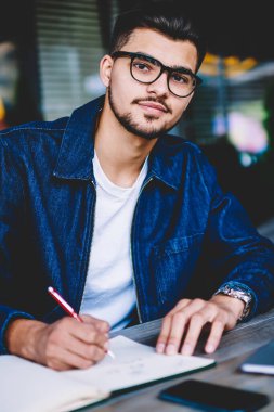Şık Eyewear online kurslar sırasında bilgileri Not Defteri'nde plan çalışma yapmaya konsantre düşünceli erkek freelancer öğrenirken not defterinde yazılı ciddi hipster adam portresi