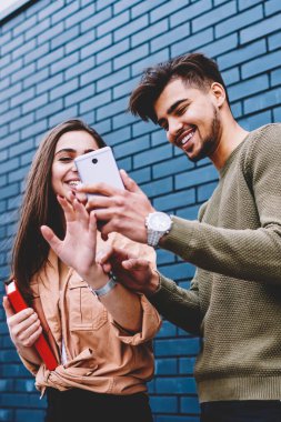 Komik fotoğraflar ile ilgilenen sosyal ağlar üzerinden smartphone dinlenme birlikte açık havada, video online tanıtım alanının yakınında telefon ayakta izleme mutlu erkek ve dişi hipsters neşeli arkadaşlarım