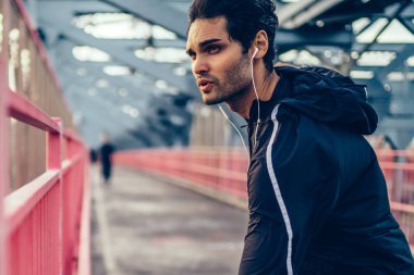 Motive fit adam kulaklık müzik mükellefi dinleme ve Koşudan önce ısınma yapıyor şık etkin elbise giymiş. Köprüde önce egzersiz germe yapıyor atletik erkek koşucu