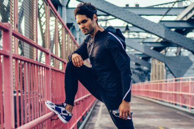 Kulaklık müzik dinleme ve eğitim modern Köprüsü'nde önce ısınma aktif giyim genç adam. Siyah eşofman ve spor ayakkabılarla şarkıları eşliğinde açık havada egzersiz sahip sportif erkekte