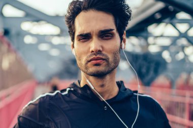 Yakışıklı sporcu portresi kamera köprüde açık havada duran modern kulaklık dinleme müzik şarkıları süre bakarak eğitim için şık eşofman giymiş. Etkin aşınmaya motive fit adam