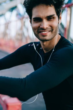 Neşeli erkek atlet portresi kameraya gülümseyen kulaklık ses dinleme siyah aktif giyim giymiş. Eşofman ayakta şişe elinde tutan kentsel ortamda pozitif sporcu