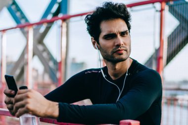 Sakallı uygun yakışıklı spor uzakta açık havada spor eğitim molası sırasında seyir kulaklık dinleme müzik hayranı. Smartphoe ve botte elinde tutan eşofman giymiş motive erkek atlet