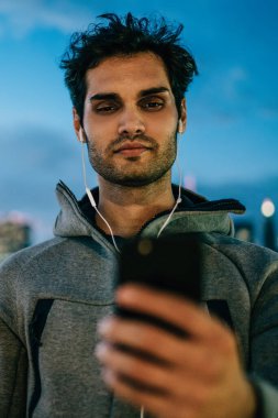 Smartphone aygıt üzerinde müzik uygulama yükleme ve açık havada akşam saat içinde kentsel ortamda ayakta modern kulaklık ses şarkıları dinleme spor ceket giymiş adam genç sakallı
