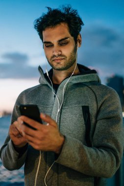 Online sosyal ağlar arkadaşlar ve dinleme radyo earphones ile mesajlaşma yakışıklı genç adam akıllı telefon açık havada akşam saat metropolis arka plan üzerinde ayakta bağlı