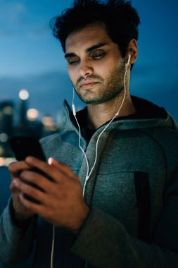 Uygulama yükleme adam genç sakallı ve ışık smartphone ekranında yansıtılır. Sakallı erkek blogger akşam kentsel ortamda hücresel ayakta bağlı earpones içinde müzik dinleme