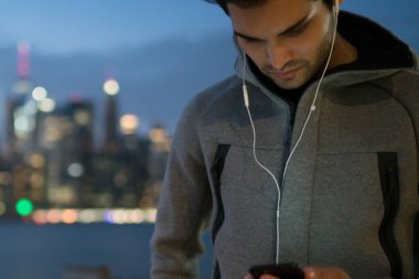 Giyinmiş genç adam spor eşofman açık havada akşam şehir arka plan ve smartphone aygıt yolu ile 4 g internet bağlantısı yükleme cool müzik ile ses çalar seçerek üzerinde duran
