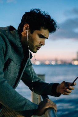 Genç adam yan görünüm çalma listesindeki modern kulaklık smartphone ayakta Kentsel şehir arka plan üzerinde akşam saat ayarında bağlı şarkıları müzik dinleme spor gri ceket giymiş