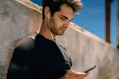 Etkin giymek için müzik çalma listesinde seçtiğiniz elinde telefon ile kentsel ortamda eğitim ayakta yan görünüm atlet giymiş. Adam akşam Player'a bağlı kulaklık ses dinleme uygun