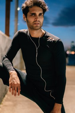 Yakışıklı uygun adam portresi kulaklık siyah eşofman dinleme müzik akşam egzersiz sırasında modern kasabada giymiş. Etkin aşınmaya geceleri bacaklarını germe yapmak motive sporcu