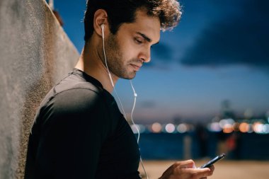 Uygun adam yan görünüm şehir promosyon arka plan kentsel ortamda çalışmadan sonra akşam üzerinde smartphone ayakta bağlı modern kulaklık müzik dinleme eşofman giymiş