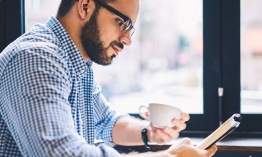 Kahve molası, yaparken defter--dan bilgi okuma raslantı giyimli hipster adam konsantre planı proje oturma pencere Kupası tutan kafeteryasında yakınındaki çalışmak için analiz erkek öğrenci