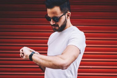 Genç hipster adam sokakta genç marka adı veya etiket, kopya alanı olan beyaz t-shirt sakallı adam saat bakarak kırmızı duvarının yakınında geziniyor buluştuğun için yürüyüş kol saati zamanında kontrol