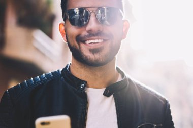 Neşeli hipster adam açık havada metin mesajları göndermek için cep telefonu kullanarak güneş gözlüğü görüntü kırpılmış, yakışıklı gülümseyen erkek sokakta yürürken smartphone cep telefonu ile online rezervasyon yapma ile memnun
