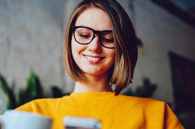 Kısa saçlı ve gözlüklü neşeli genç bir kadın akıllı telefonda komik bir bildirim okurken gülüyor. 4G interneti kullanıyor. Lezzetli kahveden ve telefonda sohbetten hoşlanan pozitif iş kadını.