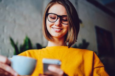 Kısa saçlı ve gözlüklü neşeli genç bir kadın akıllı telefonda komik bir bildirim okurken gülüyor. 4G interneti kullanıyor. Lezzetli kahveden ve telefonda sohbetten hoşlanan pozitif iş kadını.