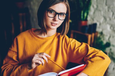 Yarım uzunluk portre çekici öğrencinin kısa saç kesimi ve şık gözlük ödev eğitim kitap okurken kameraya bakıyor. Güzel ve genç birbayan edebiyat kafede öğrenme