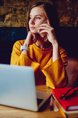 Pencereden dışarı bakan genç bir kadın kahve dükkanında dizüstü bilgisayarın başında otururken telefonla konuşuyor. Netbook 'ta uzaktan çalışma sırasında cep telefonuyla konuşan düşünceli bir iş kadını.