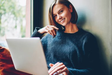 Coworking ücretsiz Wi-Fi kullanarak kendi web sitesi geliştirmek için yararlı bilgi aramak için modern dizüstü bilgisayar ile pencere üzerinde dinlenme sırasında kameraya gülümseyen güzel hippi kız portresi