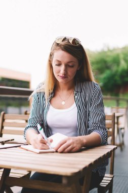 Düşünceli bir kadın en sevdiği kafeteryada eğleniyor ve deftere organizasyon planı yazıyor. Masada oturmuş ders çalışıyor.