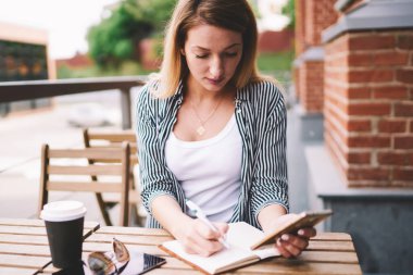 Sokak kafeteryasında okuyan konsantre bir kız öğrenci cep telefonuyla kompozisyon yazarken aynı zamanda yeni modern akıllı telefonlardan proje için hippi kız araştırma bilgileri kullanıyor.