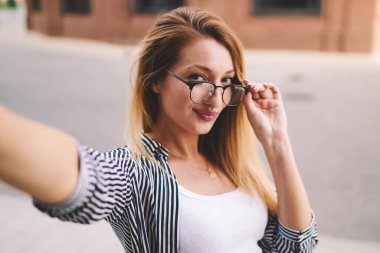 Çekici 20 'li yaşlarda, açık havada rahatlarken selfie çeken genç bir kadının yakın portresi, modaya uygun gözlük takan hippi kız şehir caddesinde yürürken kameraya bakıyor.