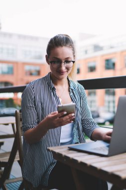 Gülümseyen hippi kız dışarıda dizüstü bilgisayarla çalışırken dikkati dağılmış, sohbet etmek için cep telefonu tutan ve netbook ile akıllı telefon multimedya dosyalarını paylaşmak için bluetooth bağlantısı kullanan kadın serbest çalışan.