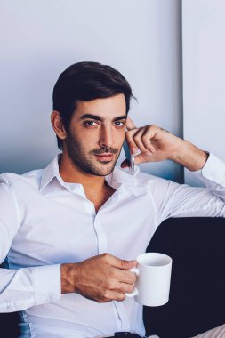 Ciddi hipster bir adam sabah kahvesinin tadını çıkarırken rahat ev koltuğunda dinleniyor ve iş görüşmeleri için meslektaşına sesleniyor. Telefon görüşmesi sırasında kameraya bakan yakışıklı bir adam.