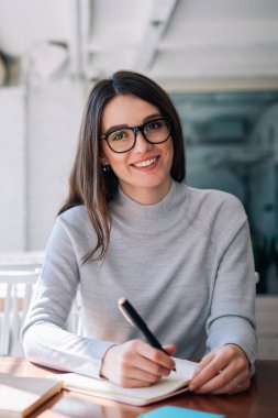 Kafeteryada eğlenen ve mola sırasında kameraya bakan gazeteci olarak çalışan neşeli hipster kız, başarılı mutlu yazar birlikte çalışma alanında otururken yeni roman fikirleri yaratarak