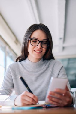 Neşeli hippi kız cep telefonuyla internete girip projeyi içeriden araştırıyor, olumlu kadın yazar akıllı telefonu tutarken makale yazıyor ve arkadaşından telefon bekliyor.