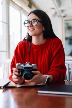 Güzel görünüşlü hippi kız eski moda teknolojiyi elinde tutarken kafeteryada hobi için boş zaman harcıyor, gözlüklü kadın fotoğrafçı evde dinleniyor.