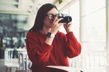 Genç pozitif esmer hippi kız üniversite kafeteryasında retro kamera ile resim çekiyor, başarılı amatör fotoğrafçı kadın odaklanmaya çalışıyor ve kapalı alanda fotoğraf çekmeye çalışıyor.