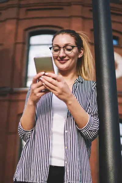 Modern elektronik cihazda bilgi arayan pozitif hipster kızı büyük binanın yanında arkadaşını beklerken; mutlu 20 'li yaşlardaki kadın gelişmiş akıllı telefondan iyi haberler alıyor.