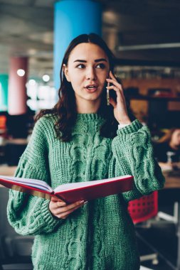 Elinde literaure kitapla dururken smartphone aygıtında iletişim meşgul esmer öğrenci. Telefon aygıtı Kitaplığı'nda ders kitabı ile cep telefonu operatörü konuşurken ile akıllı genç kadın