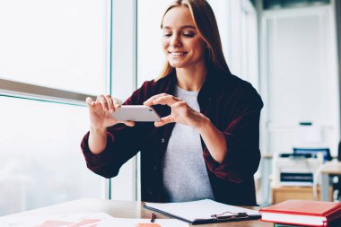 Modern Smartphone vasıl okul sırası ofiste oturan moda çizimler ve fotoğrafları yapma neşeli sarışın kadın Tasarımcısı. Cep telefonu şık studio çalışma fotoğraf çekmeye olumlu genç kadın