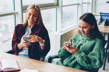 Sohbet online akıllı telefonlar üzerinde neşeli iki öğrenci Üniversitesi'nde edebiyat kitapları ile masaüstünde oturan yüksek hızlı internet bağlantısı bağlı. Pozitif hipster kızlar blogging üstünde hareket eden telefon