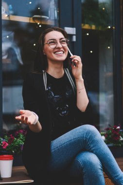 Neşeli kız öğrenci cep telefonuyla en iyi arkadaşına sesleniyor şehir ortamından memnun ve heyecanlı gözlüklü, gülümseyen hippi kız uluslararası olumlu bir konuşma yapıyor.