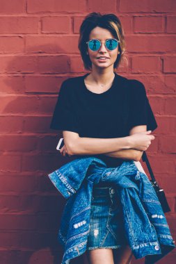 Kot pantolon ve şık güneş gözlüğü takan 20 'li yaşlardaki neşeli beyaz modellerin yarım boy portresi akıllı telefon tutuyor ve reklam mesajları ve logolar için fotokopi alanının yanında duruyor.