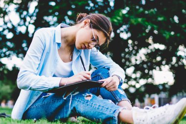 Yeşil Park, çimenlerin üzerinde boş zaman üzerinde oturan skeçler çizim çalışmaya konsantre dalgın hippi kız dinlenme sırasında proje planlama ödev görev yaparken Eyewear düşünceli kız öğrenci