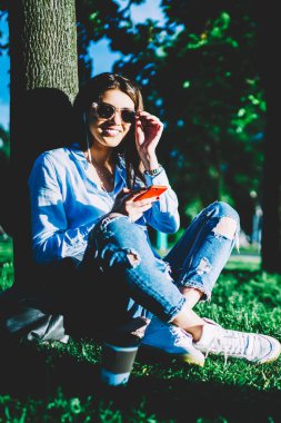 Gülümseyen hippi kız parkta uygulama mobil ve kulaklık, telefon dinleme çalma listesi şarkıları yeniden neşeli genç kadın portresi kullanarak müzik keyfi çimenlerin üzerinde oturan güneş gözlüğü