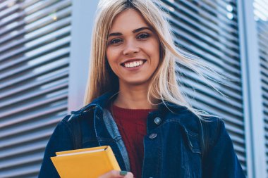 Yarım uzunlukta, yürüyüş sırasında güneşli kentsel ayarında serbest zaman zevk çekici zeki neşeli öğrenci portresi. Şirin başarılı sarışın kadın açık havada buluştuğun için beklerken kameraya bakıyor