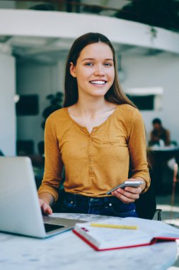 Neşeli kadın serbest çalışan kafeteryada dizüstü bilgisayarla oturup arkadaşınızı beklerken ve modern akıllı telefondan bildirimi kontrol ederken, teknolojinin gelişi ile eğleniyor.