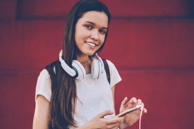 Kulaklıkla müzik dinledikten sonra kameraya bakan neşeli hipster kız portresi 4G internetle modern akıllı telefondan bildirimi kontrol ederken cep telefonuyla konuşan mutlu kadın blogcu.