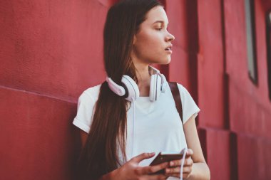 Yoğunlaşmış kadın meloman, yeni cep telefonuyla aramasını beklerken düşünceli bakışlar atıyor. Kafkasyalı kadın, modern akıllı telefonuyla duvarın yanında duruyor. Müzik uygulaması ve kulaklık için.