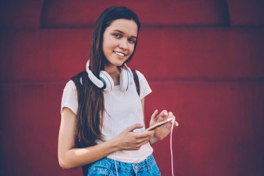 Kulaklıkla müzik dinledikten sonra kameraya bakan neşeli hipster kız portresi 4G internetle modern akıllı telefondan bildirimi kontrol ederken cep telefonuyla konuşan mutlu kadın blogcu.