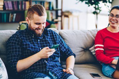 Mutlu çift modern teknolojileri ve evdeki kablosuz bağlantıyı kullanıyor. Eğlenceli, neşeli kadın dizüstü bilgisayar kullanırken erkek arkadaşı cep telefonuyla mesaj göndermeye odaklanıyor.