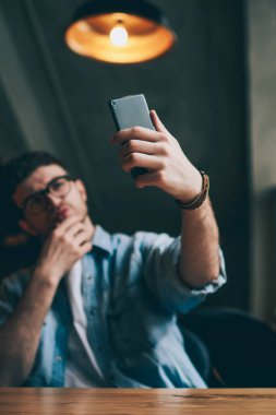 Bulanık arka planda akıllı telefon cihazıyla erkek eline seçici odaklanma, cep telefonuyla uygulamayı kullanarak sosyal ağlarda profil oluşturmak için özçekim fotoğrafları çeken genç hippi.